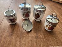 Four Royal Worcester porcelain egg coddlers in white porcelain with fruit design and metal lids on wooden surface.