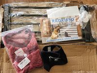 Photo of the dog gate parts in packaging, a pink small dog sweater labeled size XS, and a black dog harness labeled size XXL, all laid out on cardboard.
