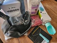 Photo shows gray and black YUDODO dog carrier, pink dog sweater, Aqua blue portable dog water bottle with attached bowl, black hair comb, and opened package of dog diapers on wooden floor.