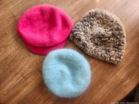 Top view of three soft fuzzy hats: bright pink beret, light blue fuzzy hat, and brown/grey textured hat.