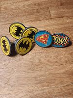Top view of six superhero themed drawer pulls arranged on wood surface showing four Batman logos, one Superman logo, and one POW! logo