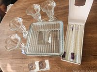 Clear vintage square glass refrigerator dish with lid, four flared-top ice cream sundae glasses, boxed taper candles, and two glass candle holders arranged on wooden table.