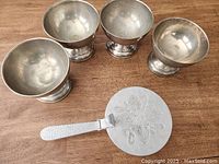Four nickel silver bowls arranged in a rough square on wooden table, slightly tarnished interior, next to aluminum flip-top pan with white textured handle and floral engraved lid.