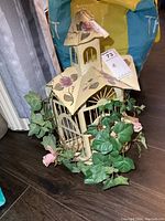 Front and side view of cream-colored metal decorative bird cage with floral painted roof and artificial green vine with pink flowers wrapped around base.