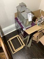 Photo showing two tall grey metal lantern battery candles on top of a boxed Mr. Coffee appliance, with three wooden frames leaning against the box, and a small wooden key rack.