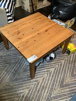 Photo showing the square wood coffee table on a patterned rug next to other household items.