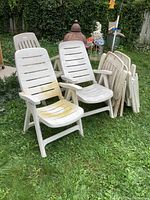 Set of six white resin folding deck chairs arranged on grass, two chairs open in front with visible dirt and discoloration, four folded behind, showing two different slat patterns.
