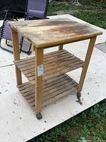 Full view of the solid wood kitchen island cart showing the top surface with stains and wear, middle and bottom slatted shelves, and caster wheels.