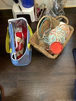 Photo showing a wicker basket containing bulky pastel-colored yarn, knitting needles, embroidery hoops, and a red cylinder container, next to an insulated bag holding red and white yarn skeins and more knitting needles.
