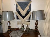 Photo showing both matching table lamps with wooden bases and silver lampshades on a wooden table, with wall hanging behind.