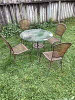Photo showing round glass top metal frame patio table with four metal frame chairs with bamboo slats around it on grass.