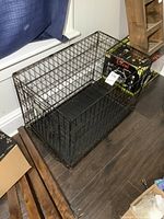 Side view of black metal wire pet kennel with a plastic tray inside, placed on dark wooden floor next to a window.