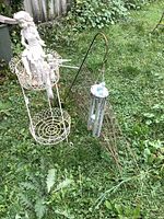 Photo shows white metal plant stand with resin angel seated on top, metal wind chime hanging from hook, and tomato cages laid on grass.