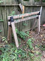 Pair of large wooden sawhorses standing outdoors in front of a wooden fence, showing full view and overall condition.