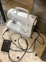 White Singer Tradition sewing machine on floor with power cord and foot pedal, taken from a side angle showing machine controls and handle.