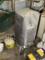 Gas smoker with cover and propane tank in a storage area with dirt floor, some rust and dust visible on cooker