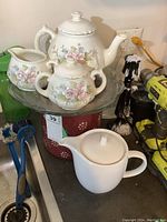 Full view of floral ceramic teapot, creamer, sugar bowl on glass tray and white teapot on counter, with large ceramic canister in background