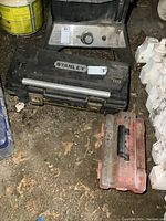 Black Stanley 26-inch toolbox placed on a dirty floor, alongside a smaller red plastic case likely containing hole saws and drill bits.
