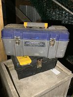 Two toolboxes stacked: a blue and gray Mastercraft plastic toolbox with yellow handle on top, black metal toolbox below with yellow organizer box on top