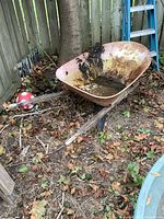 Side angle showing the rusty bowl of the wheelbarrow, dirt inside, wooden handles visible, and flat tire on the ground.