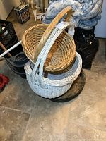 Three wicker baskets stacked together, one painted white with large handle, two natural color with different handle styles, on a tiled floor.