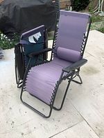 Two folding zero gravity chairs shown, one open and one folded, purple fabric seating with black metal frame and armrests, with lumbar pillows present.