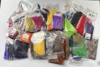 Top-down image showing an assortment of approximately 60 small plastic bags containing colored feathers for fly tying, including colors purple, yellow, green, red, orange, black, and natural tones.