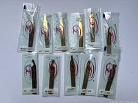 Photo showing 11 individually packaged Renzetti fly fishing tying tweezers in gold and dark metal tones laid out on a white background.