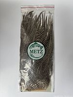Front of sealed clear plastic package showing multiple striped rooster hackle feathers, Metz label in center with green trout logo.
