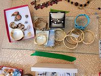 Top view of costume jewelry including several gold-tone bangles, blue and gold-tone bracelets, and multiple pairs of coral-colored earrings arranged on a glass surface.
