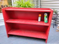 Front view of the red painted wood bookcase on an outdoor deck, showing two main shelves and an upper shallow shelf, with visible scratches and scuffs.