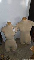 Front view of female and male fabric-covered clothing display forms on concrete floor, showing beige fabric and wooden neck caps.