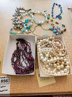 Overview of assorted costume jewelry including beaded necklaces and bracelets displayed on textured surface with two small boxes holding jewelry pieces
