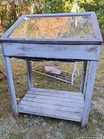 Front view of a weathered wooden greenhouse box with glass top panel and lower shelf.