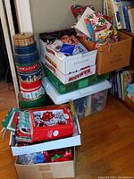 Wide view showing stacked holiday themed decorative metal cans, boxes of gift bags, ribbons, and bows clustered on floor next to bookshelves.
