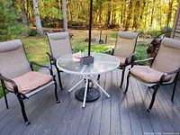 Four outdoor patio chairs arranged around a glass top metal table on a wooden deck, each with a cushion on the seat. Background is a wooded yard.
