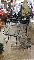 Two metal plant stands with floral design, rectangular shape, black metal frames and weathering visible, large wood birdhouse shaped like a church with painted finish. Also metal frame side table with glass top and several wire baskets in various sizes and shapes sitting around on the ground in a garage setting.