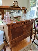 Front angle view of wood side board with mirrored back, glassware on top, and clock. Shows three small drawers with wooden knobs, large cupboard with sliding doors, and curved legs.