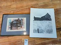 Photo of two signed prints on floor: one framed color house print and one black and white barn print with plastic cover