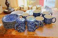 Six dark blue spatterware mugs and multiple blue and white patterned saucers and plates on wood surface.