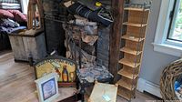 Photo shows a living area corner with a tall wicker and metal standing shelf, two footstools, culinary painted plaque, wood hanging shelf, and other decorative pieces arranged near a stone fireplace and window.