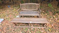Outdoor teak bench and corresponding teak table on fallen leaves with visible weathering and natural patina.