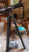 Tasco model 302059 black telescope mounted on a black tripod with adjustable legs in a room with boxes and books.