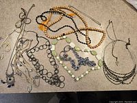 Overhead view of assorted jewelry items spread on beige carpet, showing wooden bead necklace, black bead strands, metal chains, and green oval bead necklace.