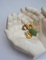 Matte white cupped hands sculpture holding several pieces of jewelry, including a green stone pendant, gold hoops, and a ring.