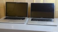 Photo showing two Apple MacBook Pro laptops side by side, lids open with screens off and keyboards visible