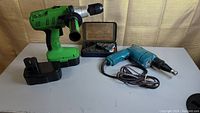 Green cordless drill with side handle and battery pack, blue corded drill, and metal box with drill bits arranged on a table.