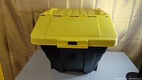 Front view of a black plastic storage bin with a yellow hinged lid closed.