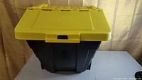 Stackable black plastic storage bin with a hinged yellow lid closed, showing the front and lid latch.