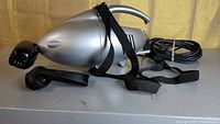 Silver portable vacuum with brush attachment and strap, showing the full unit with power cord.